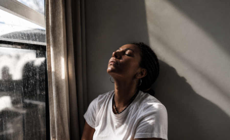 Woman sitting beside a window with her eyes closed as sunlight falls across her face, suggesting reflection and patient trust.