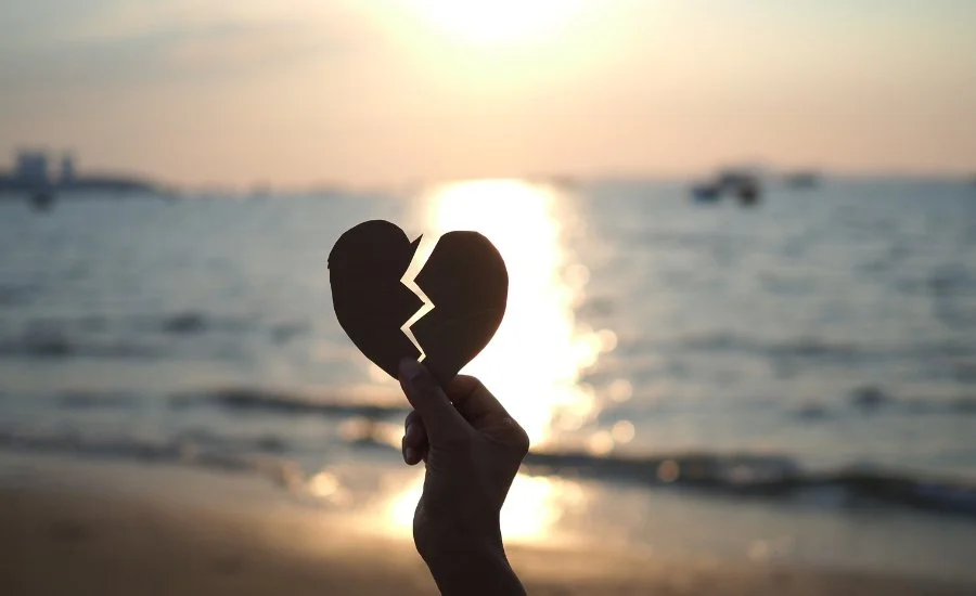 A hand holds up a paper heart torn in two, silhouetted against a sunset over the ocean.