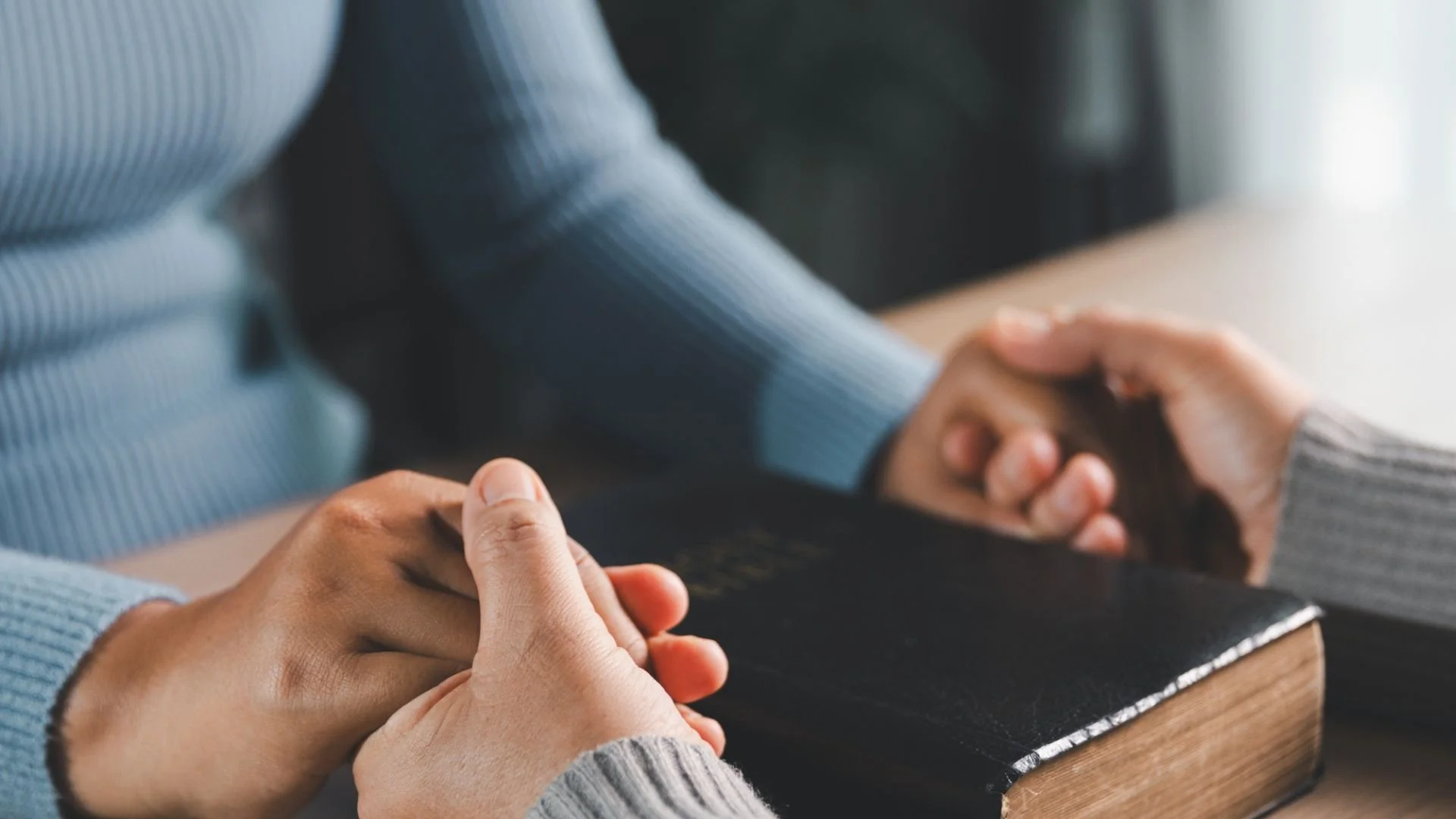 Two people holding hands praying with a Black Bible between them on a table