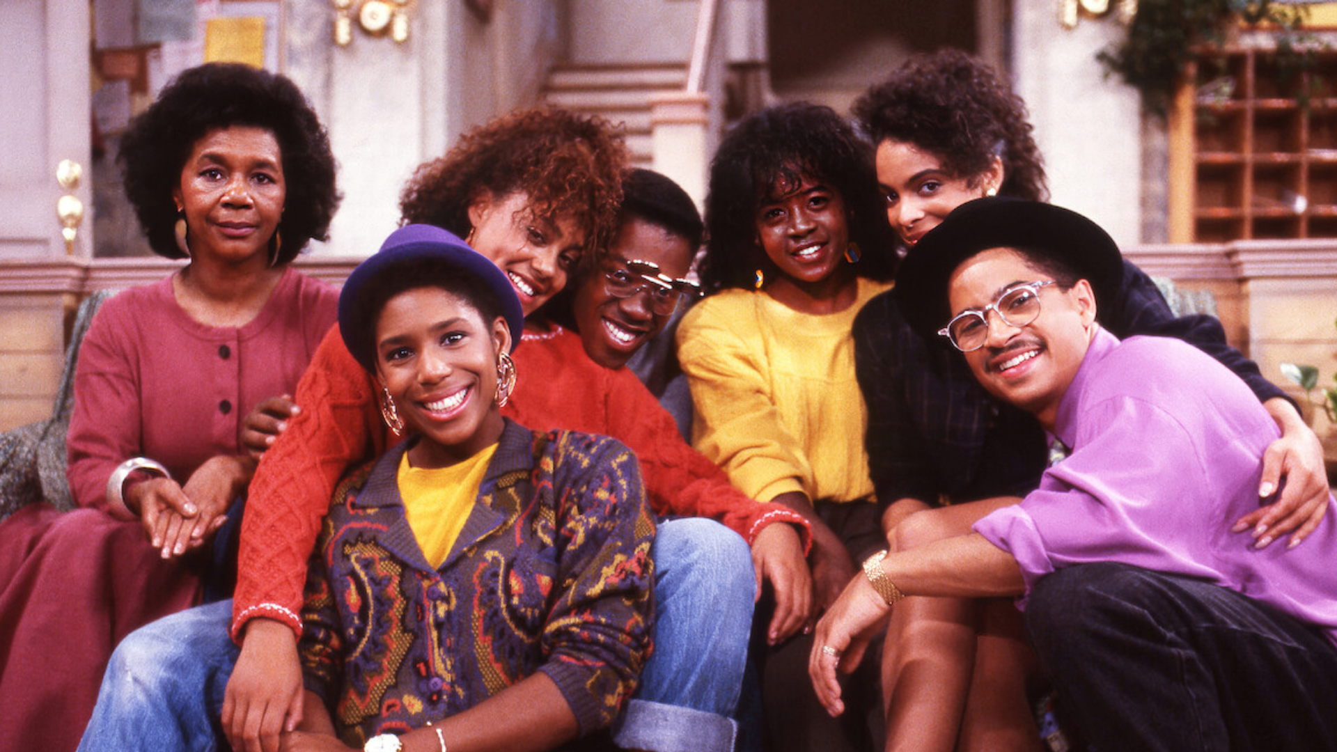 Seven cast members of A Different World pose together on the Hillman College dormitory set, smiling and leaning on one another in colorful late-1980s fashion, including bold sweaters, wide-brim hats and oversized glasses.