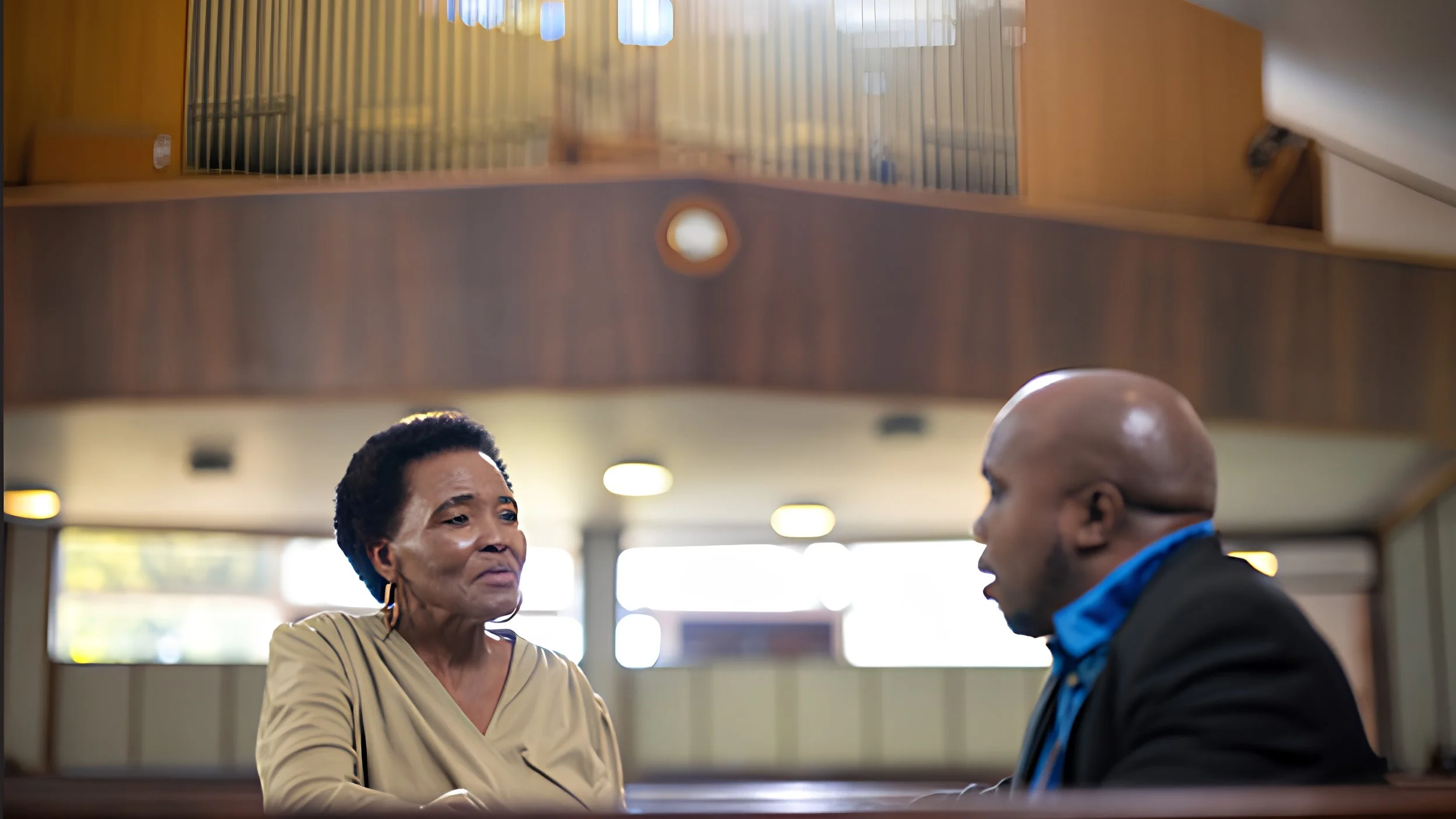 An older woman in a tan top listens as a man in a dark suit jacket and blue shirt speaks to her while they sit in church pews, with organ pipes and a balcony behind them.