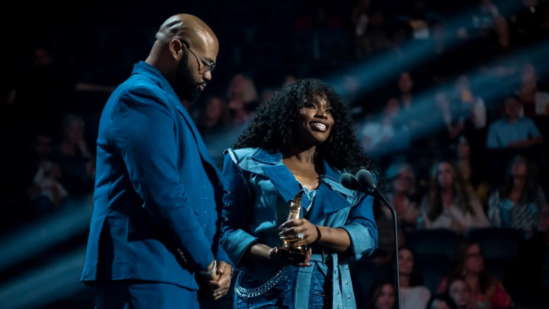 Tasha Cobbs Leonard, in a blue denim ensemble, smiles while holding a Dove Award statuette at a microphone as her husband, Kenneth Leonard Jr., in a blue suit, stands beside her with head bowed