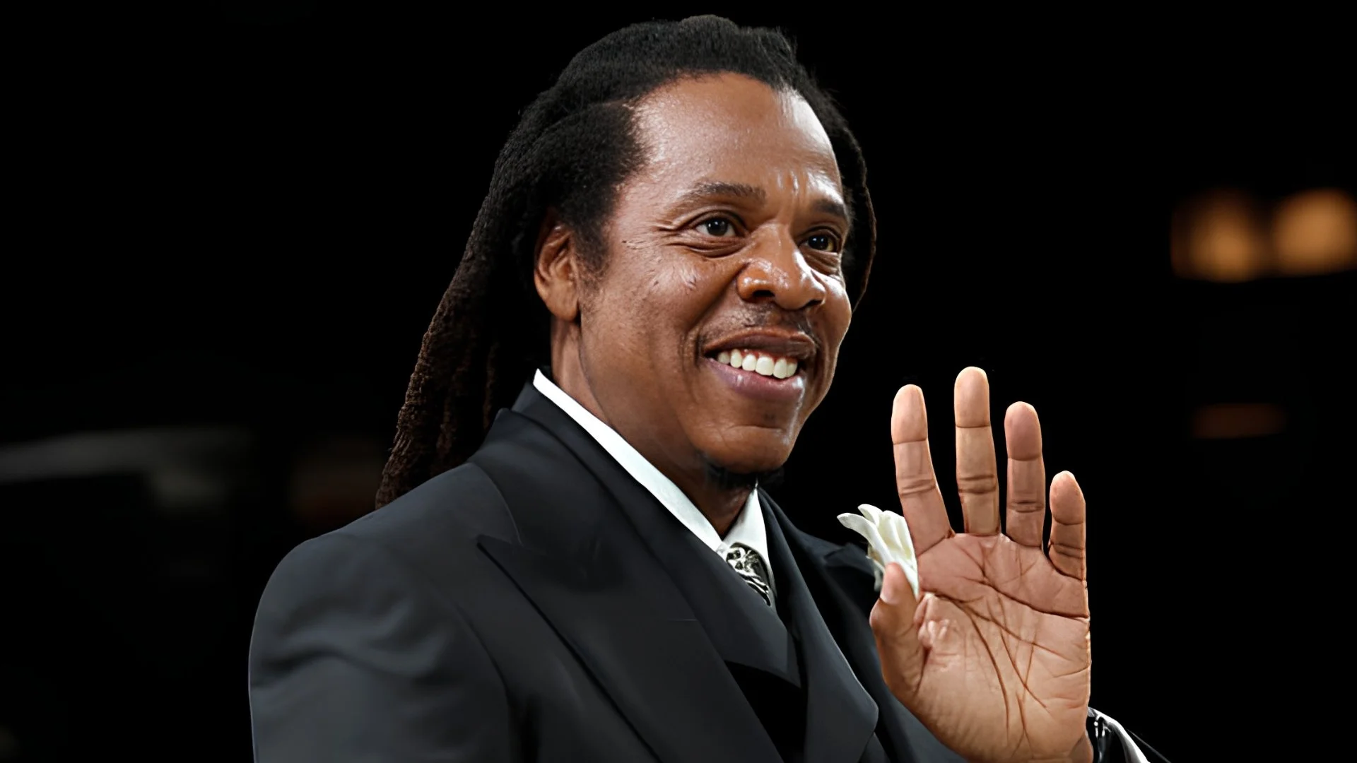 Jay-Z, wearing a black suit with a white flower on the lapel, smiles and waves.