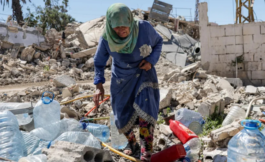 An elderly woman in a blue dress and green hijab walks through concrete rubble and scattered water containers near a destroyed building in southern Lebanon.