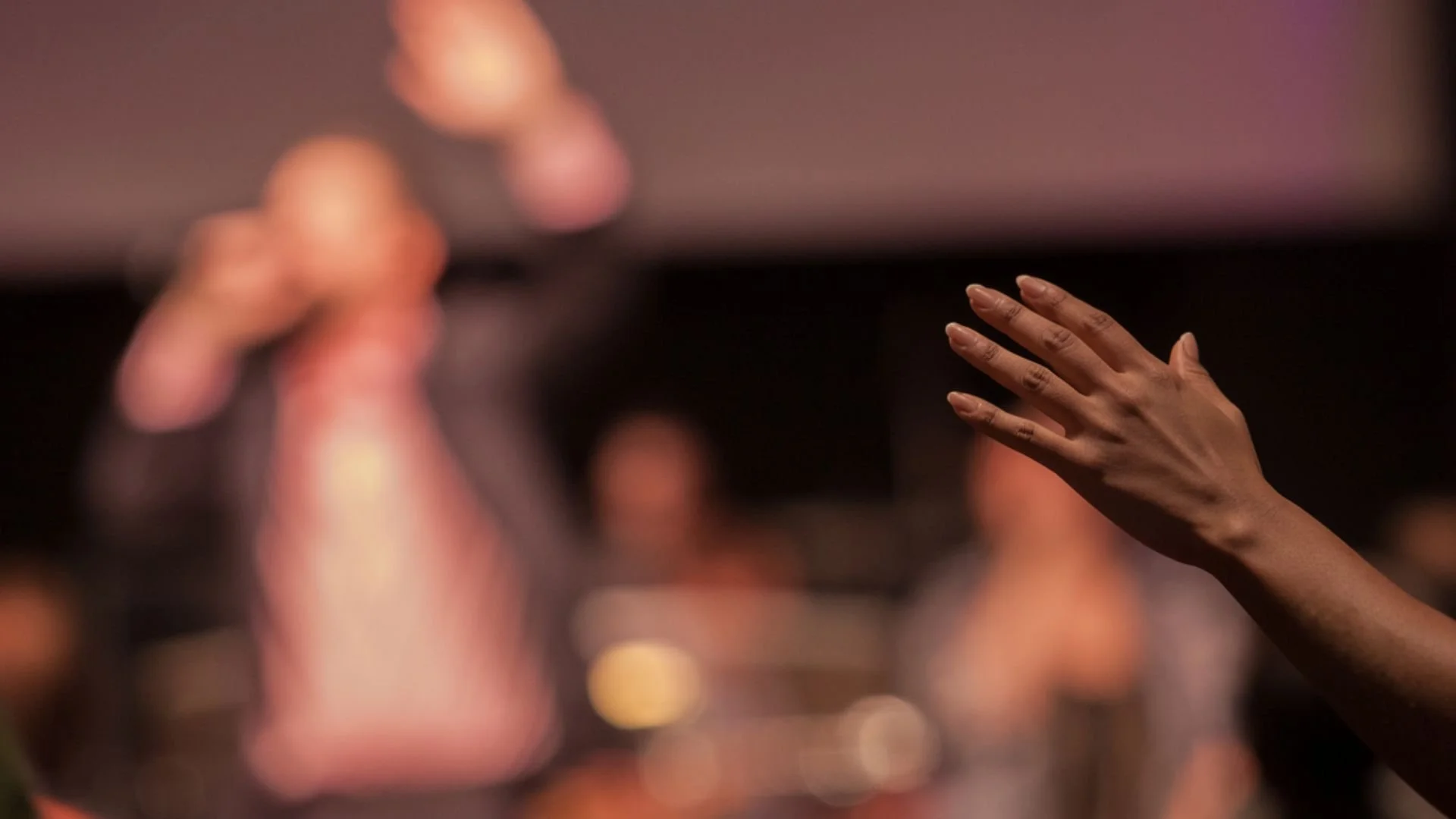A worshiper’s raised hand in the foreground as a blurred leader holds a microphone on a church stage.