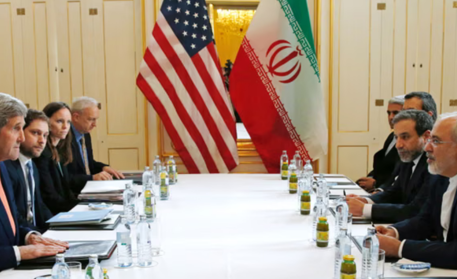 U.S. and Iranian officials sit across from one another at a negotiating table in Vienna, with American and Iranian flags displayed prominently in the background.