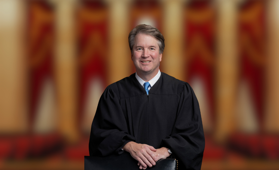 Official portrait of U.S. Supreme Court Justice Brett Kavanaugh wearing a black judicial robe, seated with his hands folded before a blurred interior background.