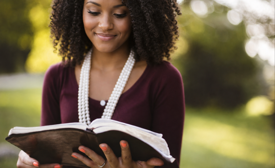 A Black woman reading an open Bible outdoors, smiling gently