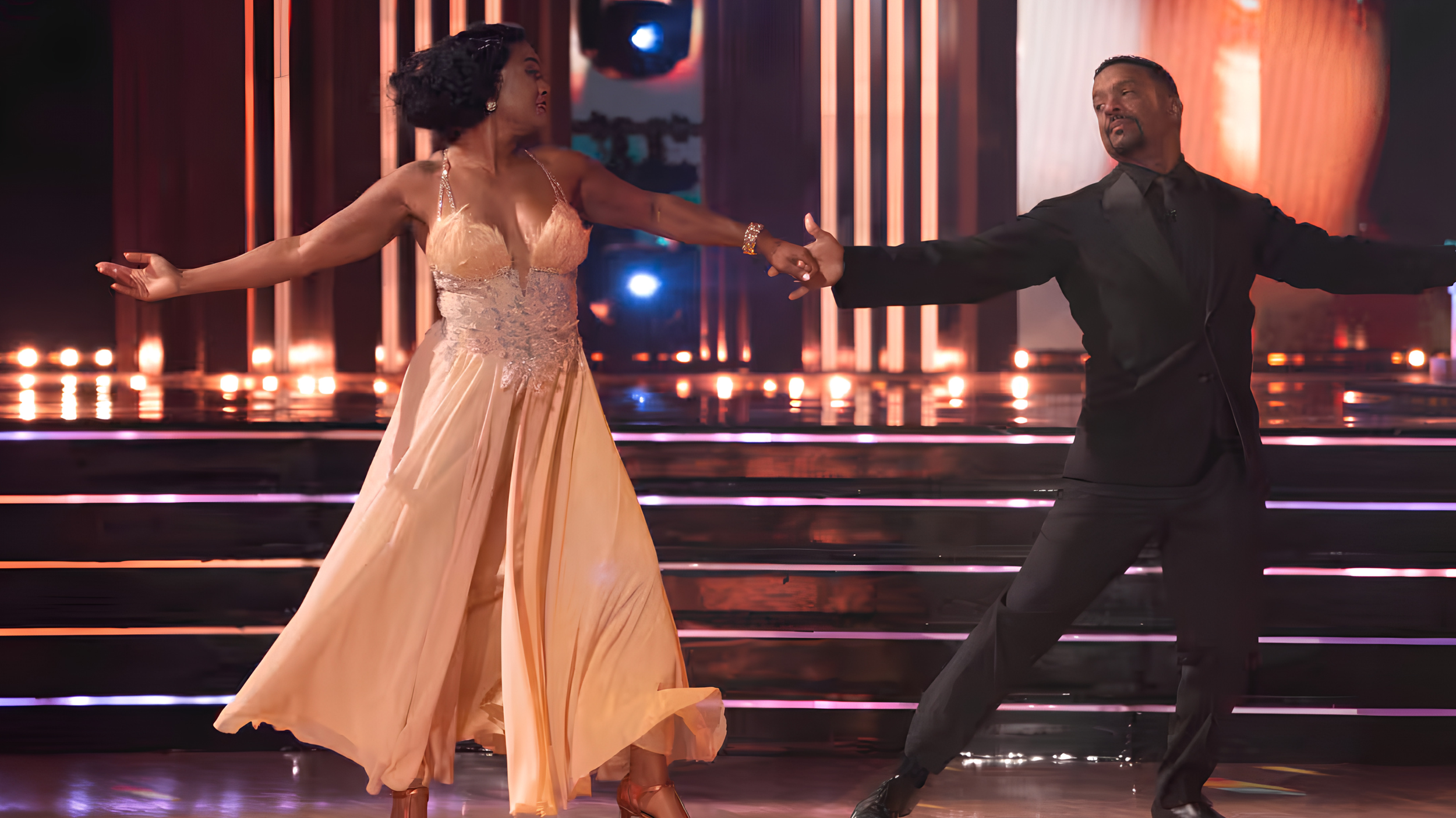 Tatyana Ali in a flowing peach gown with a feathered bodice dances hand in hand with Alfonso Ribeiro, dressed in a black suit, across the Dancing With the Stars ballroom floor under warm stage lights.