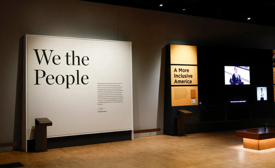 Interior of the Obama Presidential Center museum tower showing a large "We the People" wall display alongside an "A More Inclusive America" exhibit with video screens and wooden seating