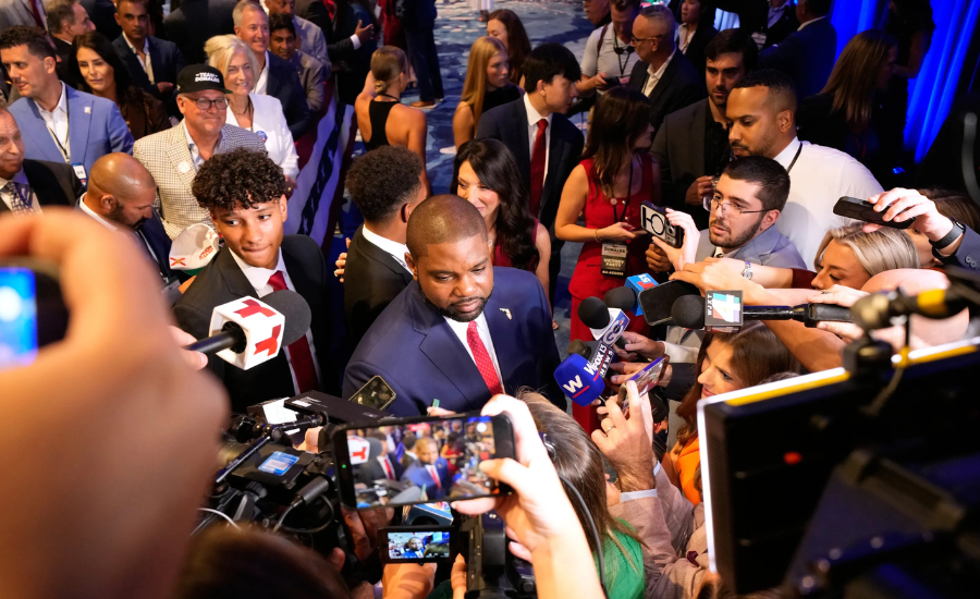 Byron Donalds, surrounded by reporters holding microphones and phones, speaks to the press amid a crowd of onlookers.