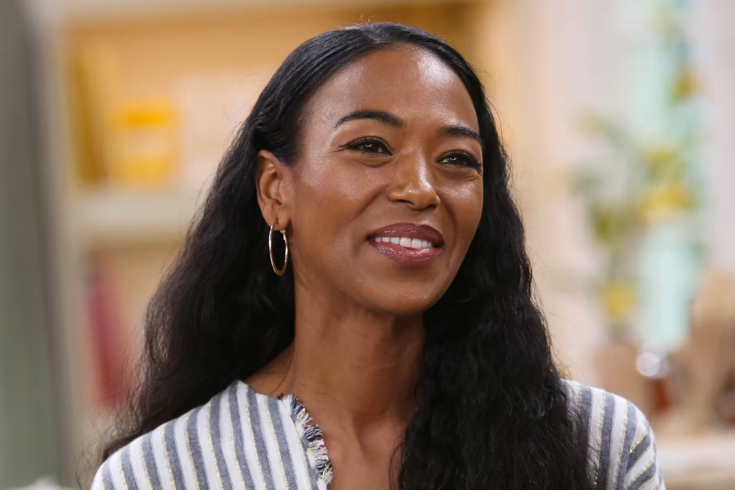 Ananda Lewis smiles during a 2019 television appearance, wearing gold hoop earrings and a gray and white striped top, her long dark hair falling past her shoulders.