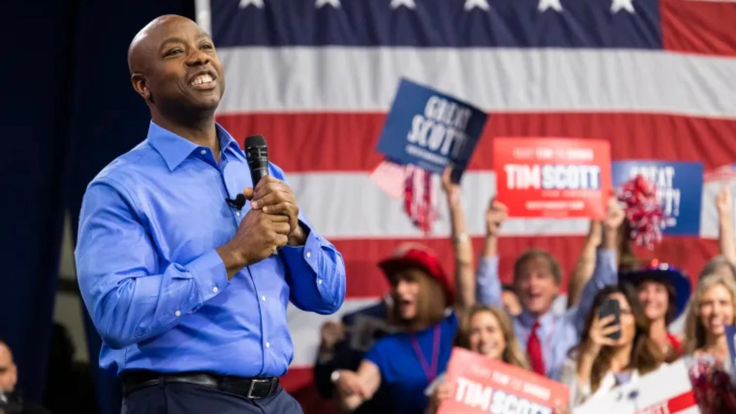 Tim Scott, in a blue dress shirt and holding a microphone, smiles on stage in front of a large American flag as cheering supporters wave “Tim Scott” and “Great Scott!” signs.