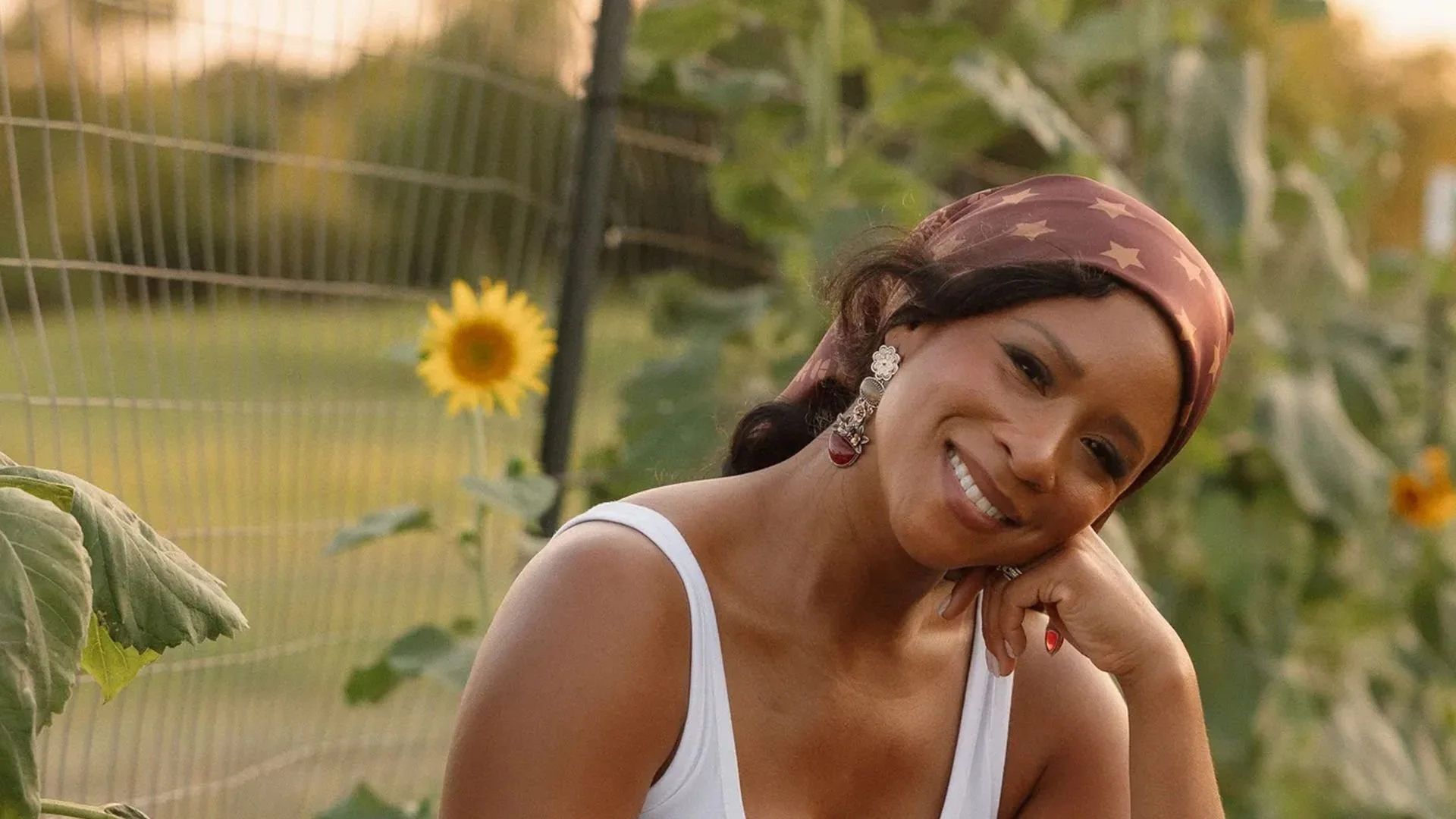 Aisha Patterson, wearing a white tank top and a patterned headscarf, smiles with her head resting on her hand in a garden with sunflowers behind her.