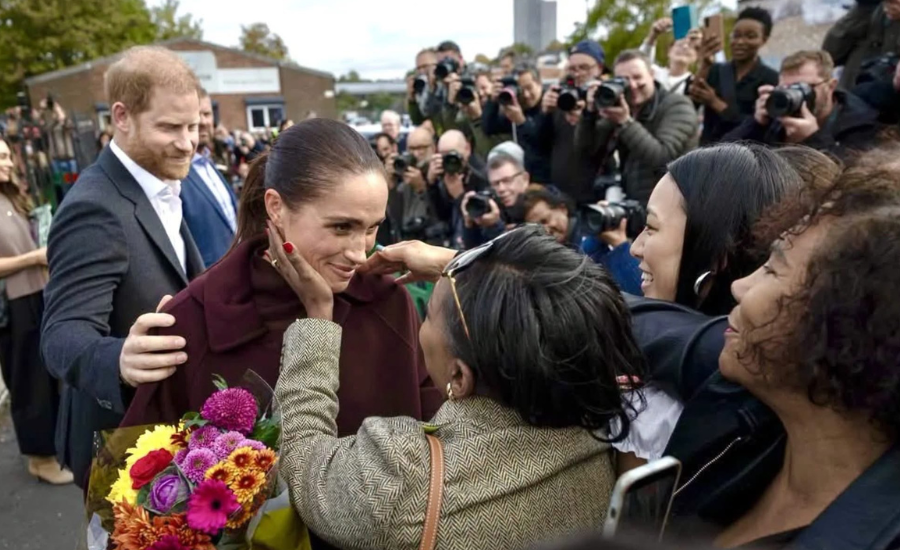A woman reaches up to hold Meghan's face as Meghan, holding a bouquet of bright flowers, smiles; Harry stands at her side and a line of photographers aims cameras at them.