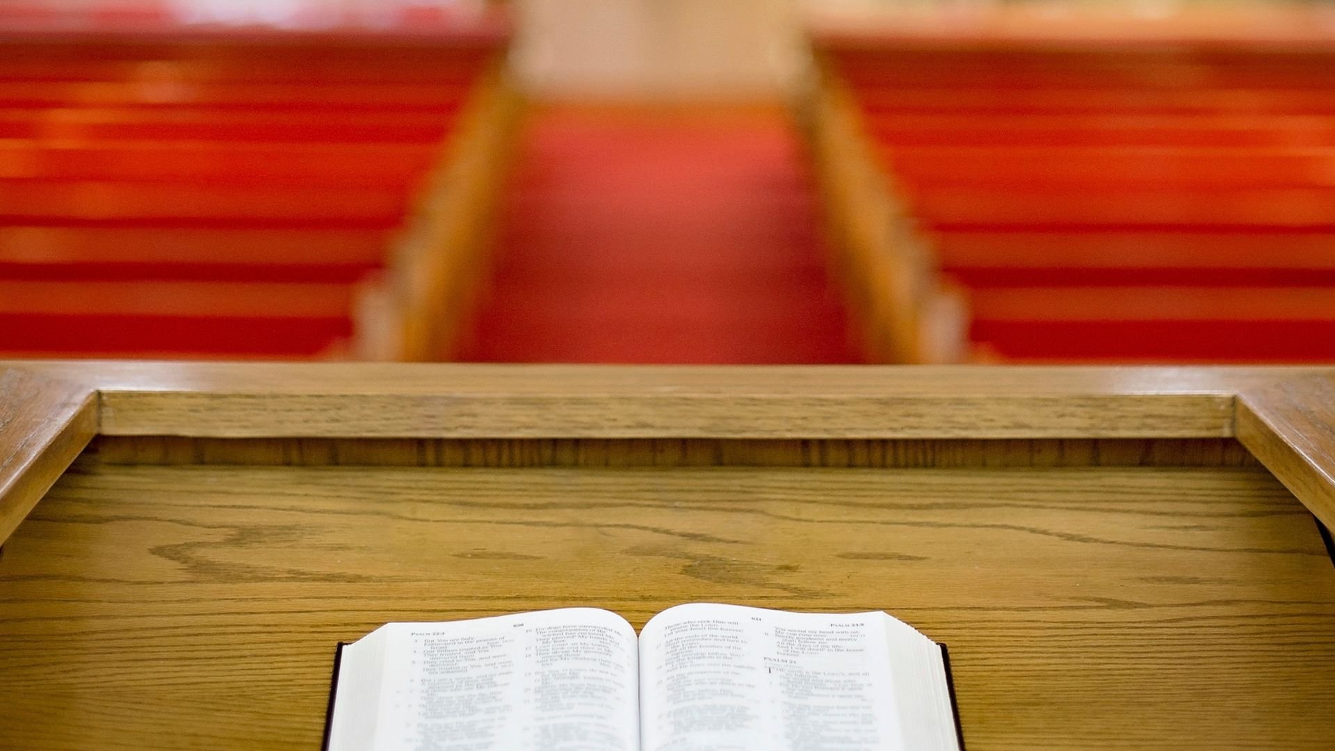Open Bible on a wooden pulpit facing empty red-cushioned church pews and a center aisle.