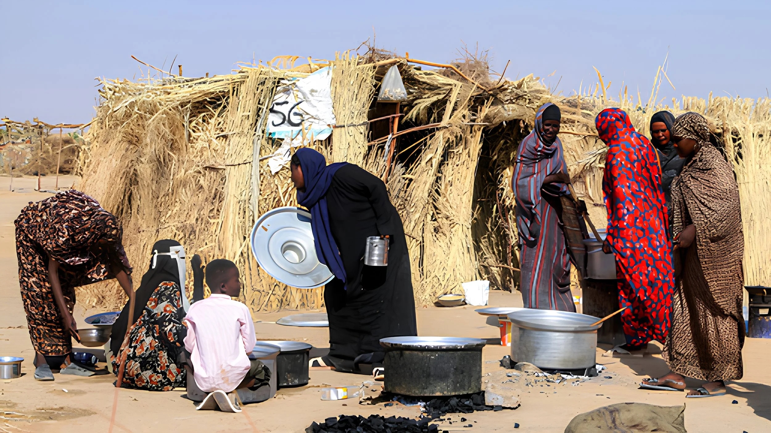 Women in long patterned dresses and head coverings cook in large metal pots over charcoal outside a thatched shelter, as a boy in a pink shirt sits nearby.