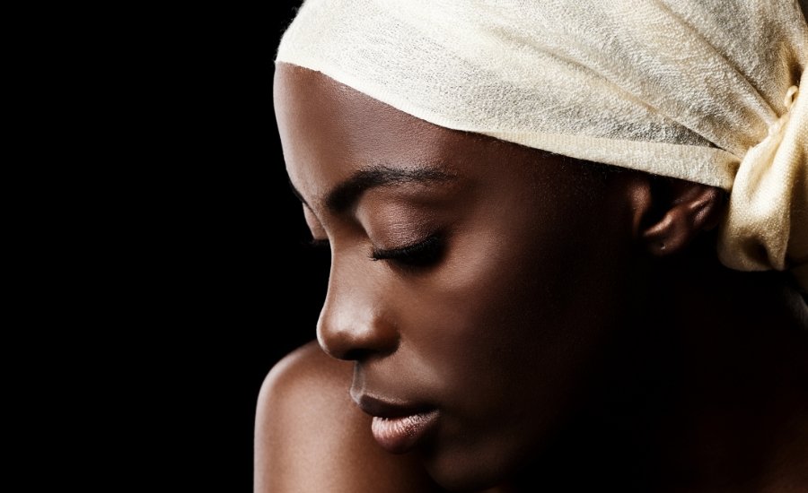 A Black woman wearing a cream head wrap bows her head with her eyes closed against a dark background.