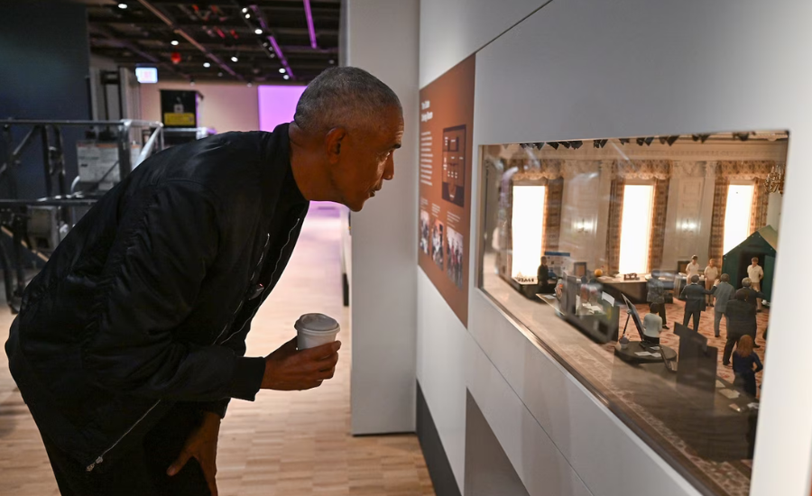 Former President Barack Obama leans forward holding a coffee cup to look closely at a miniature diorama exhibit inside the Obama Presidential Center museum