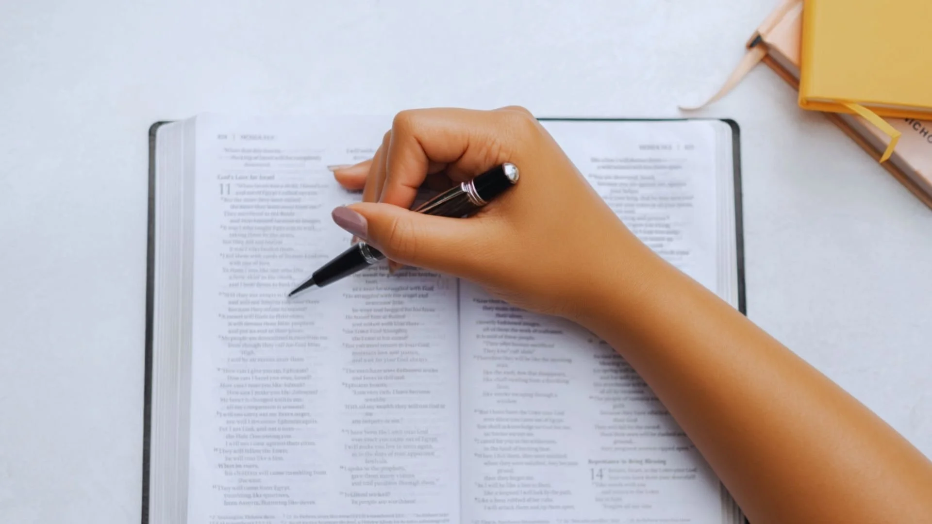 A woman’s hand holding a pen over an open Bible on a white table, with journals nearby.