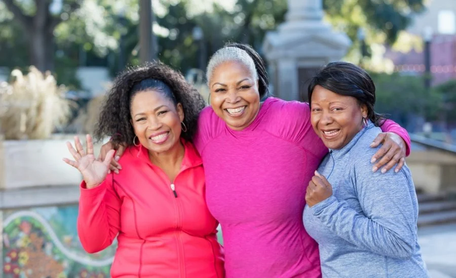 Three smiling Black women in athletic jackets stand arm in arm in a city plaza.