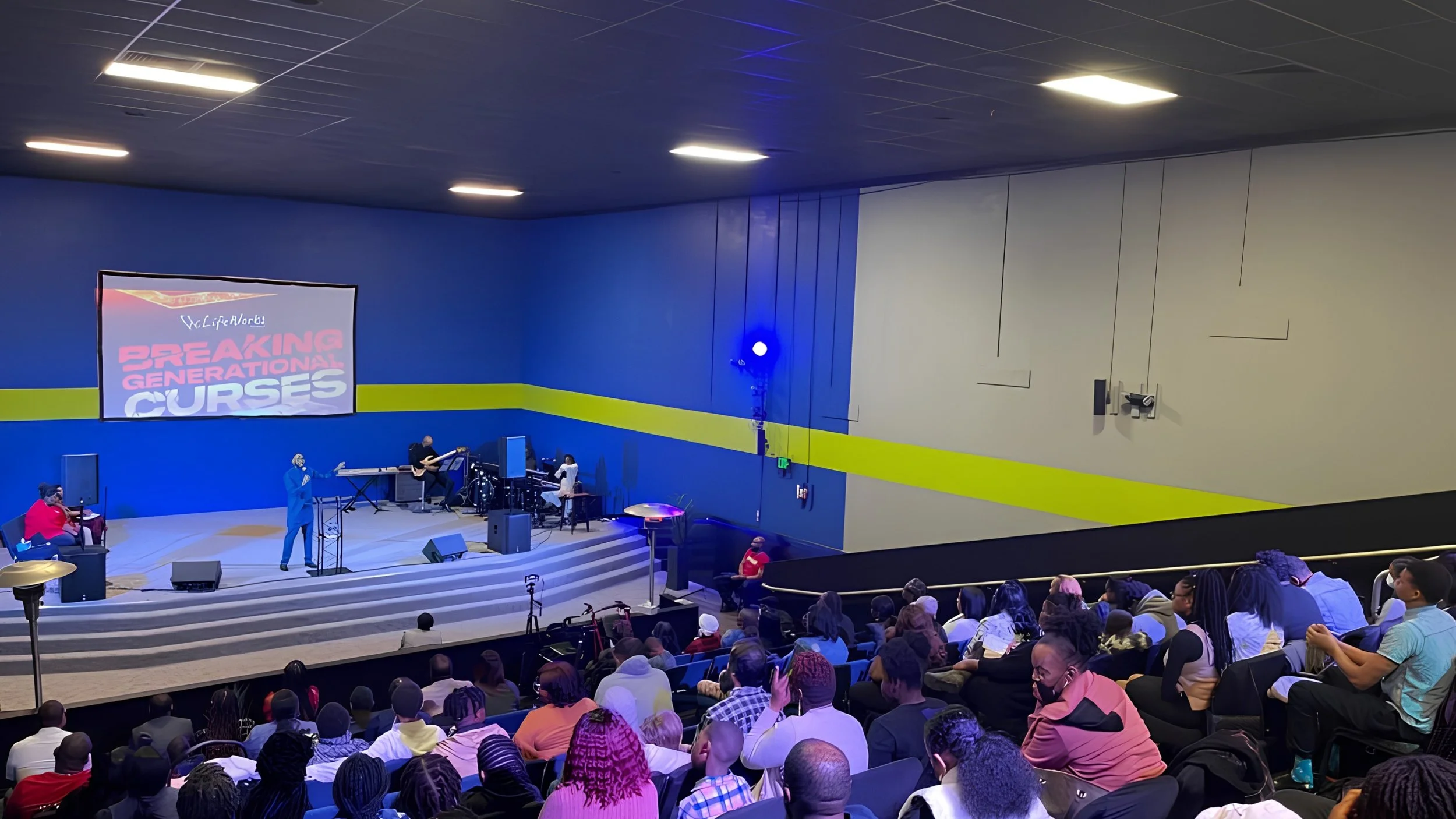 A preacher in a blue suit speaks from a stage at LifeWorks Church while musicians play behind him. A screen above the stage reads “Breaking Generational Curses,” and a congregation fills the seats in a sanctuary with blue and lime-green walls.