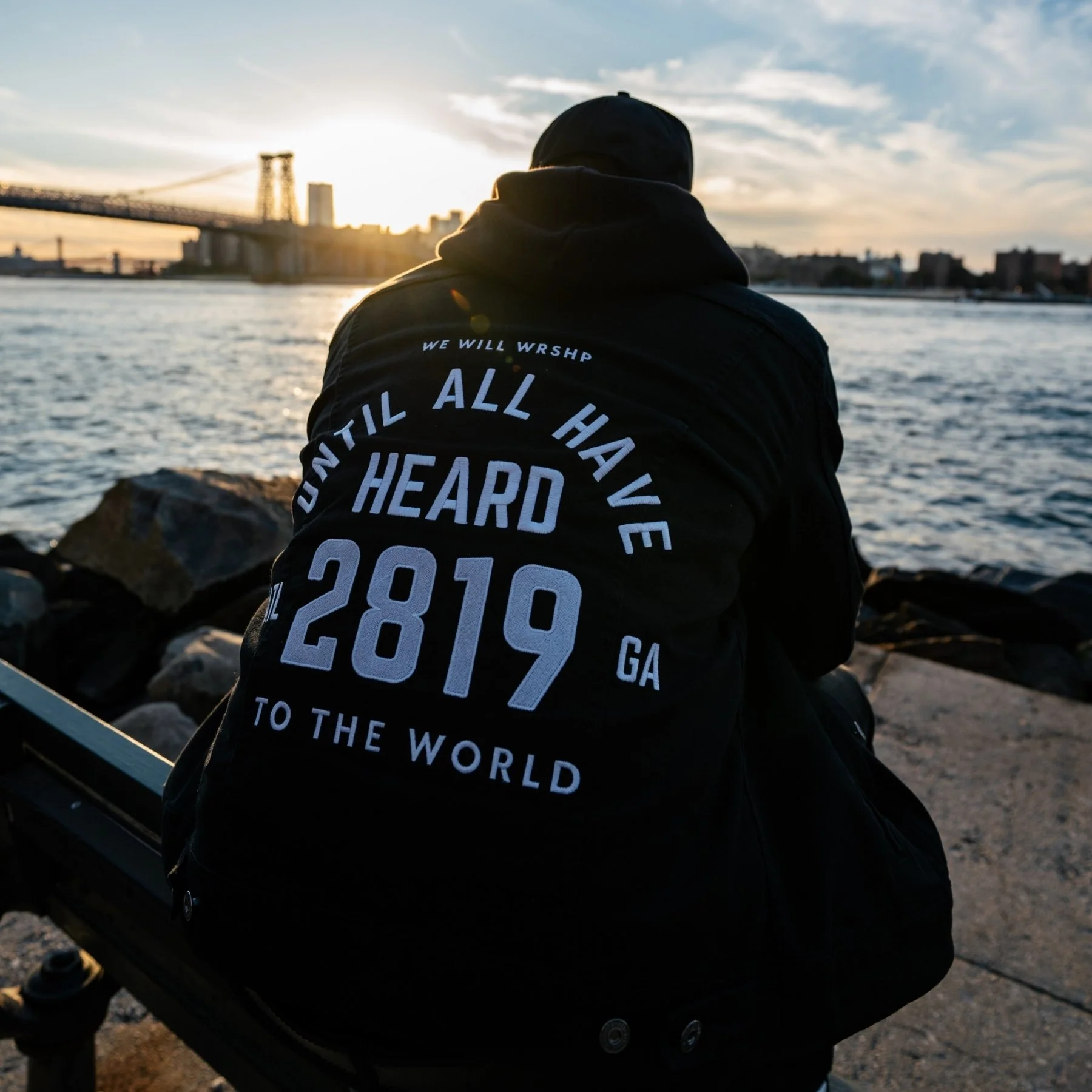 Pastor Philip Anthony Mitchell, seen from behind, sits on a waterfront railing at sunset facing a bridge and city skyline. The back of his black jacket reads “We Will Wrshp, Until All Have Heard, ATL 2819 GA, To the World.”