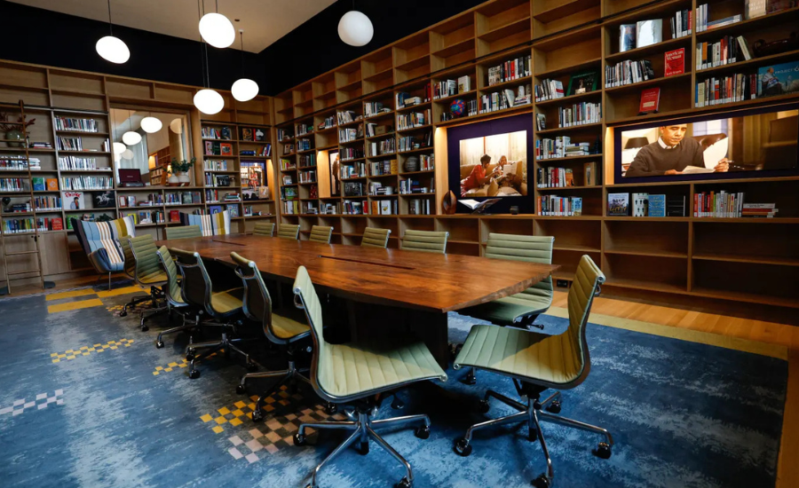 A conference table surrounded by chairs inside the Obama Presidential Center branch of the Chicago Public Library, with floor-to-ceiling wooden bookshelves, photographs, and globe pendant lights