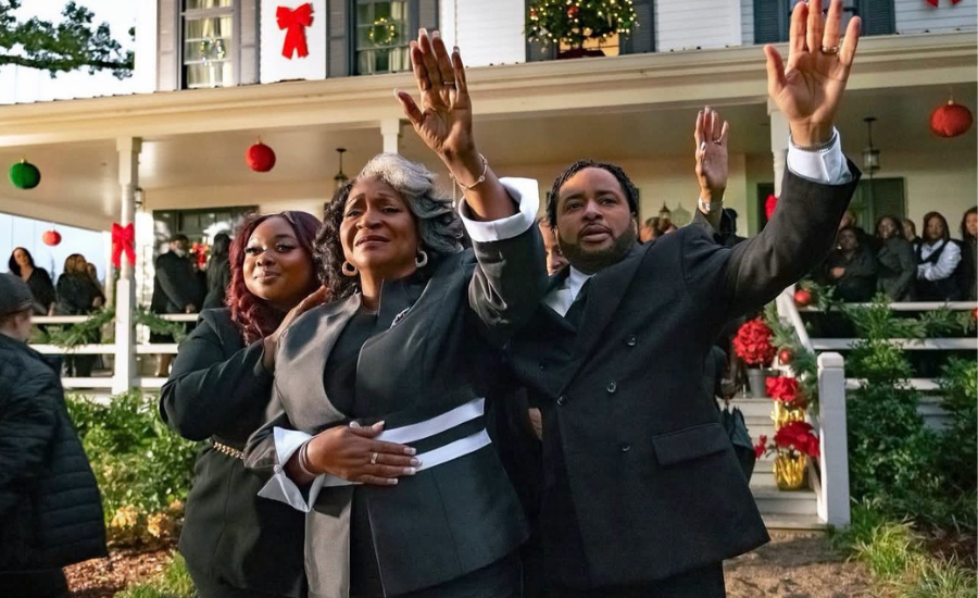 Three people in dark formal clothing raise their hands in praise outside a white house decorated for Christmas as a crowd watches from the porch.