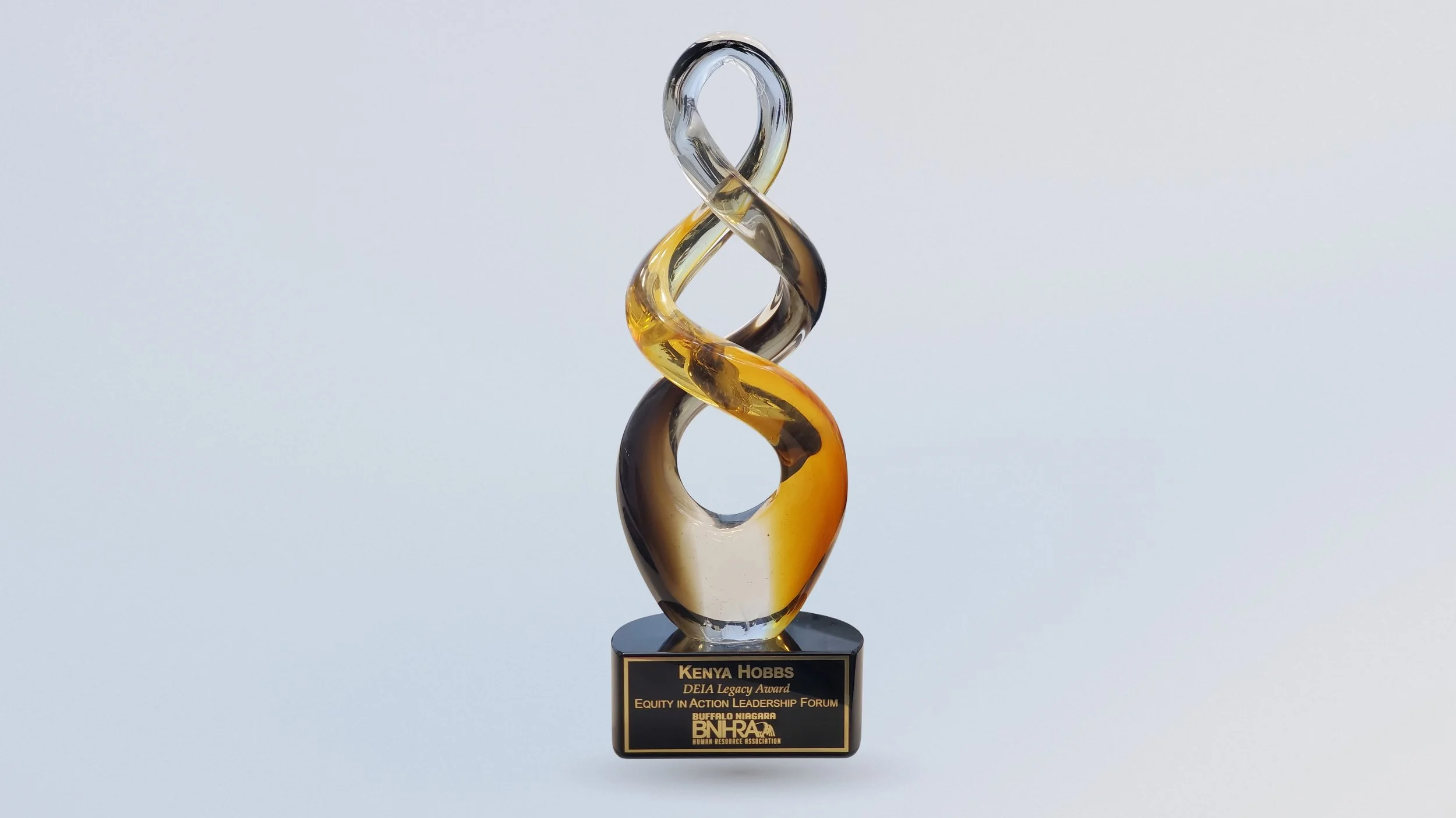 A twisted glass sculpture in amber, clear and smoky tones rises from a black base engraved with “Kenya Hobbs, DEIA Legacy Award, Equity in Action Leadership Forum” and the Buffalo Niagara Human Resource Association logo.