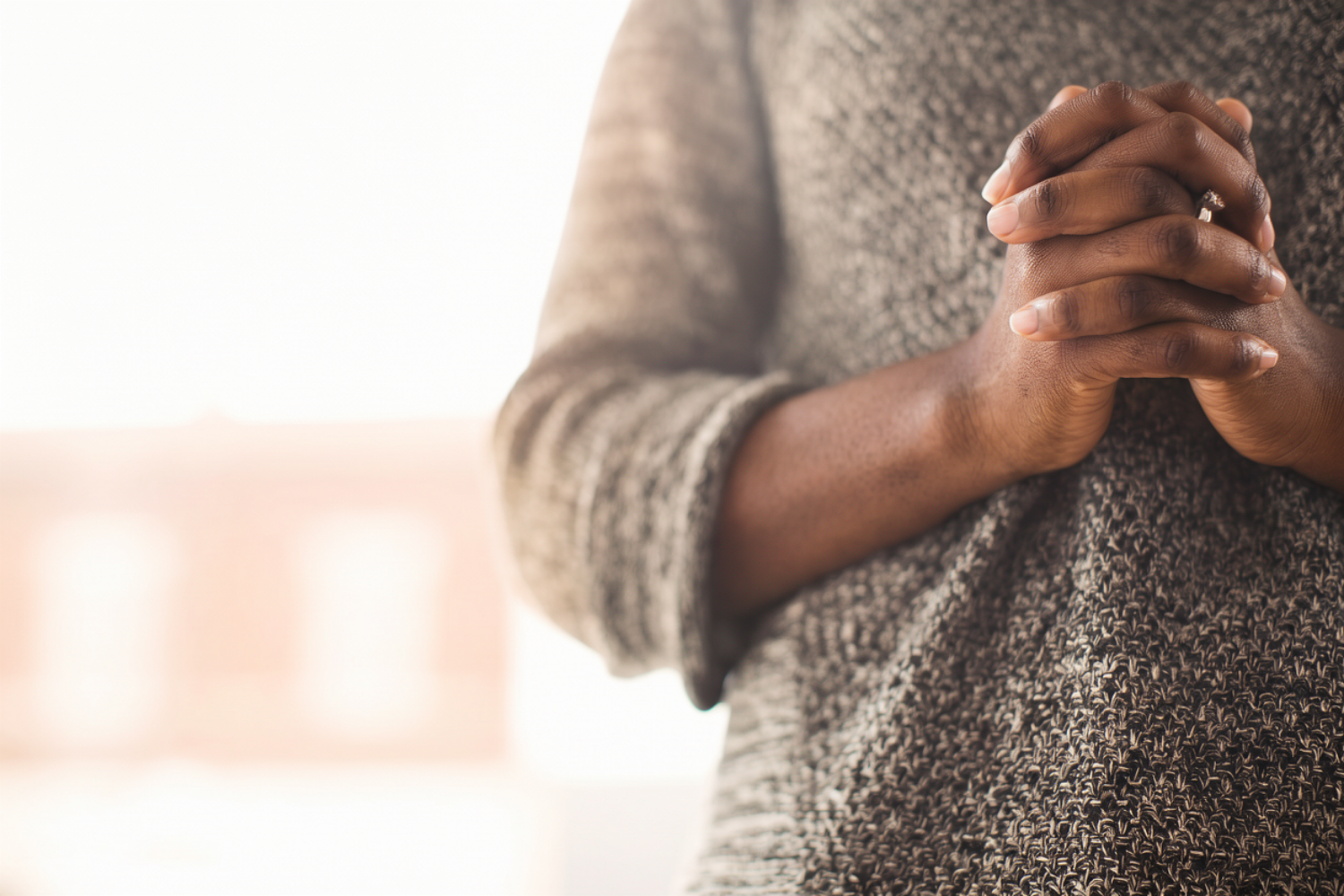 Close-up of a Black woman’s hands clasped in prayer against a gray knit sweater, with soft window light glowing in the background.