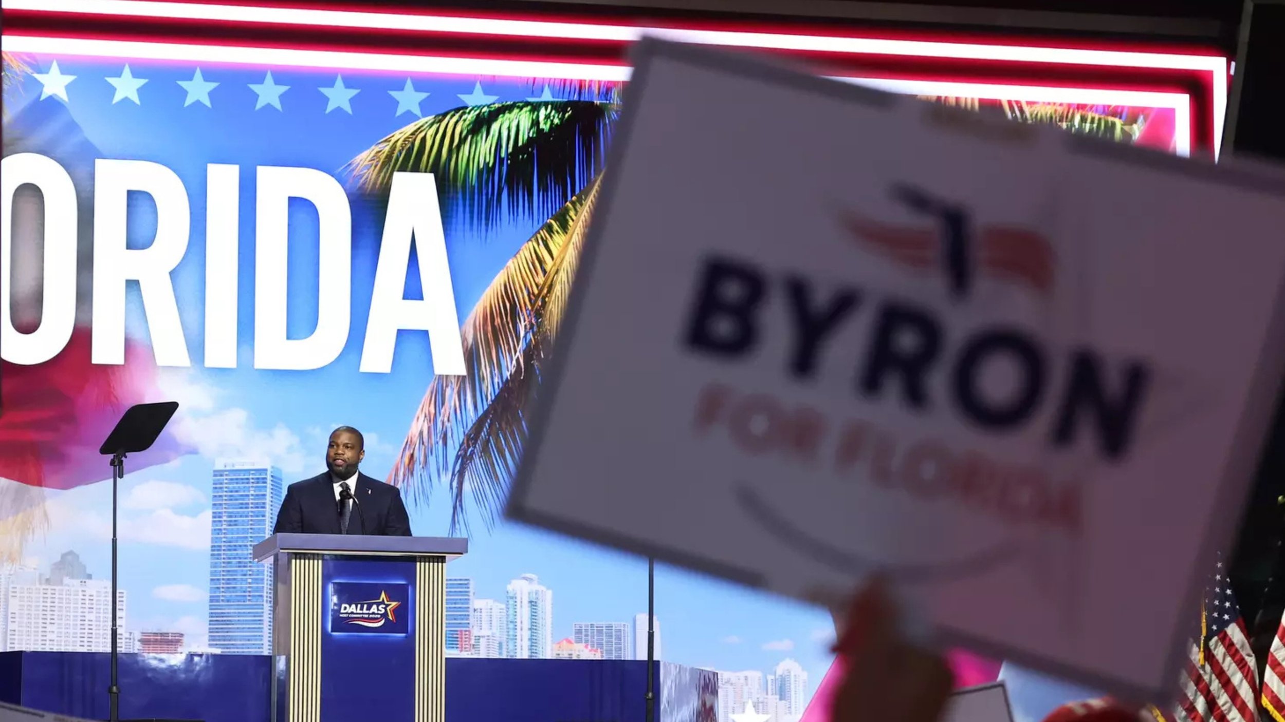 Byron Donalds speaks from a podium marked “Dallas” in front of a large screen reading “Florida” with palm trees and a city skyline, while a supporter in the foreground holds a “Byron for Florida” sign.