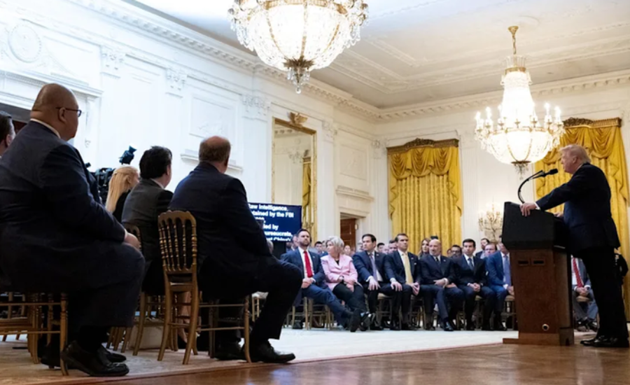 President Trump speaking at a podium before a seated audience in the White House East Room, with a screen displaying intelligence-related text in the background.