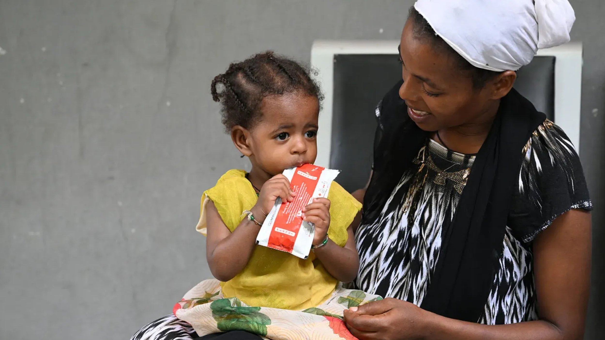 A small child in a yellow dress holds a red and white sachet to her mouth as a smiling woman in a white head wrap looks on.