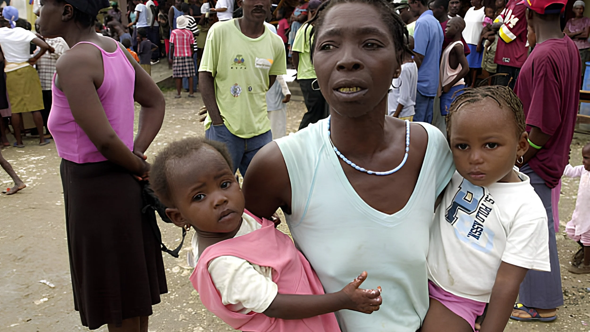 A woman in a light blue top holds a toddler on each hip as a crowd gathers behind her on a dirt lot.