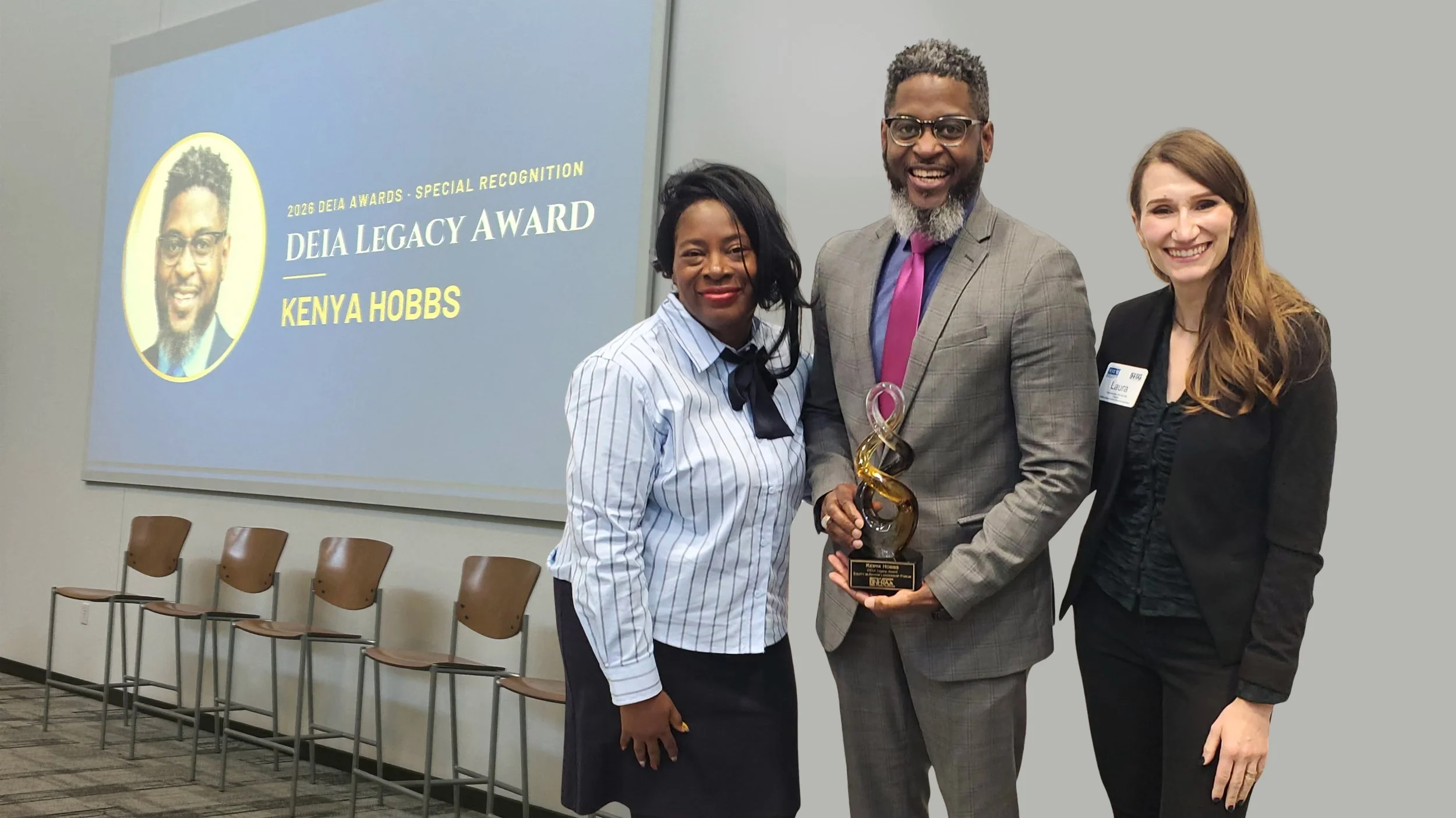Kenya Hobbs holds a glass DEIA Legacy Award between LaTanya Walton-Brown and Laura Azzarella, beside a screen announcing him as the 2026 DEIA Legacy Award recipient
