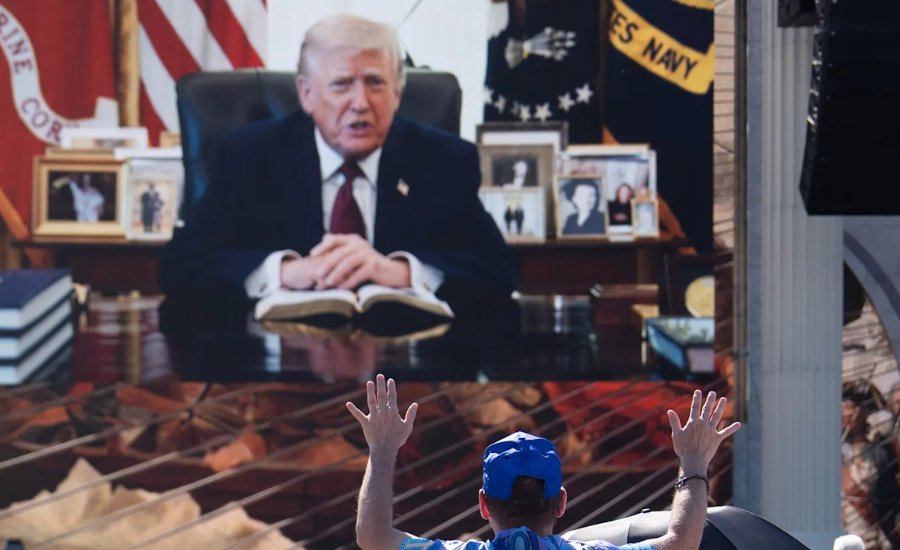 Attendee with hands raised in worship facing a large screen showing a pre-recorded video message from President Donald Trump seated at a desk with an open Bible during the Rededicate 250 event on the National Mall on May 17 2026