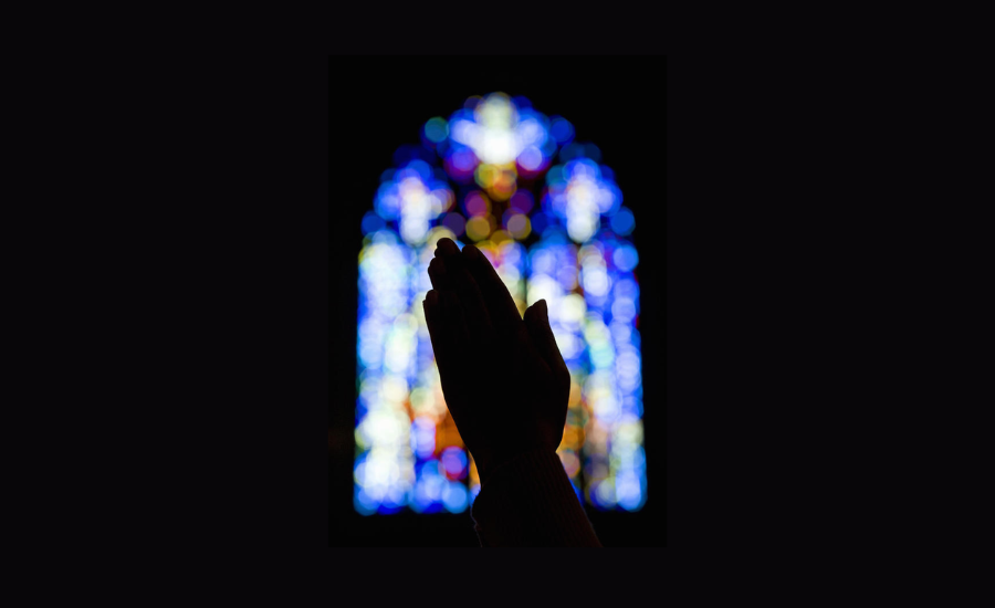 Silhouette of two hands pressed together in prayer in front of a blurred stained glass church window glowing with blue and multicolored light against a dark background
