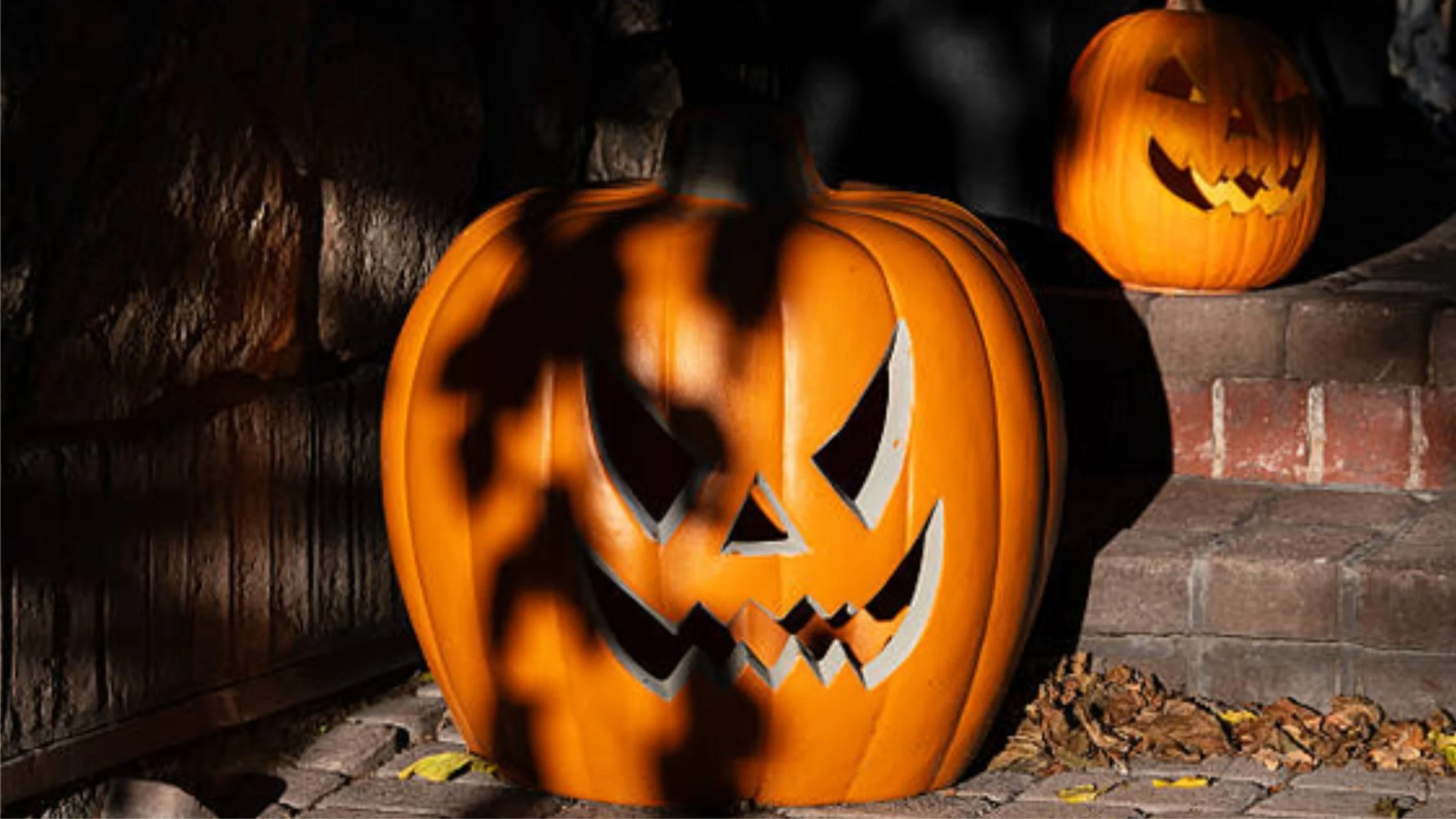 Two orange plastic jack-o’-lanterns with jagged, menacing carved faces sit on brick porch steps at night, partly shadowed by leaves, with dry fallen leaves at their base.
