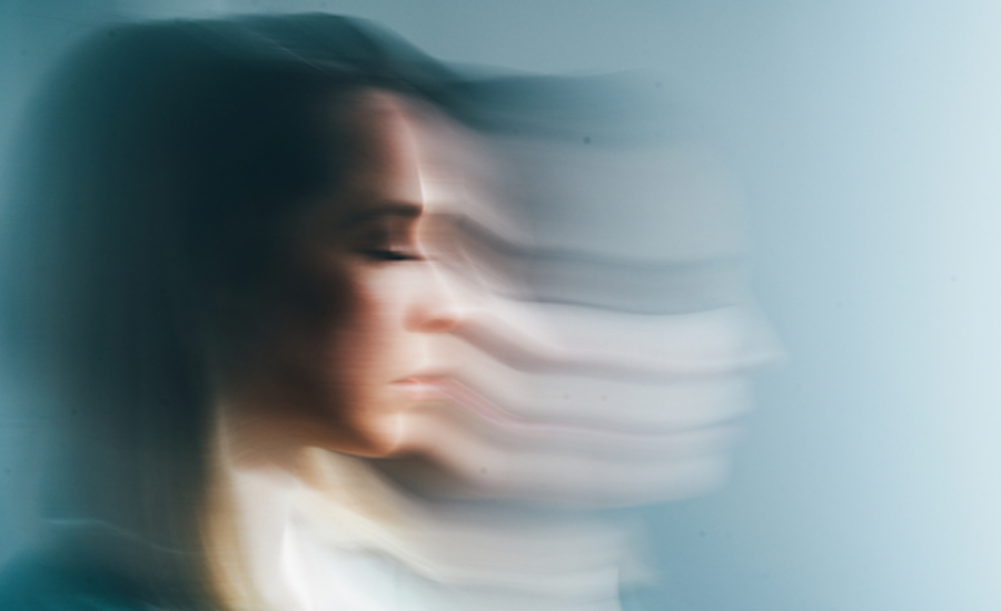 A woman's face in profile with hair swept across it, photographed with motion blur against a soft blue-gray background.