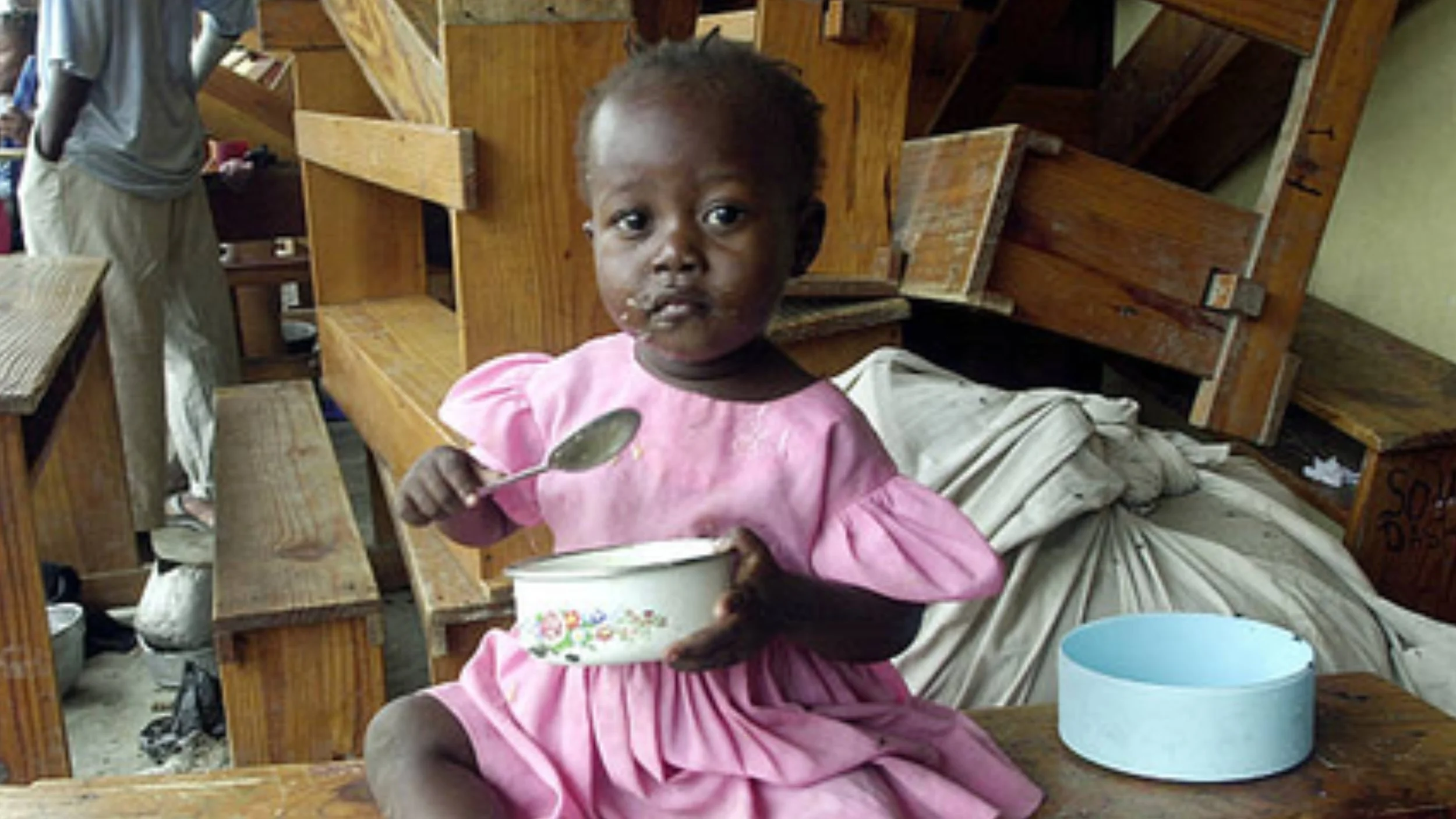 A young girl in a pink dress sits on a wooden bench holding a bowl and spoon, with stacked school desks and bundled belongings behind her.