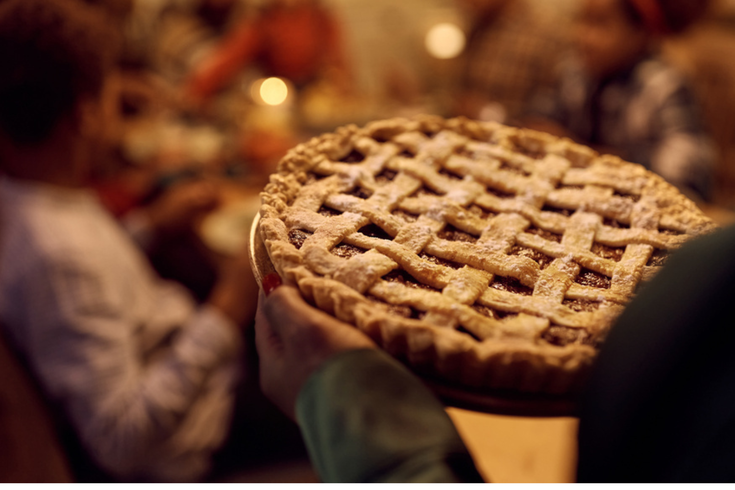 Hands carrying a lattice-top pie dusted with powdered sugar toward a candlelit Thanksgiving table where people are gathered