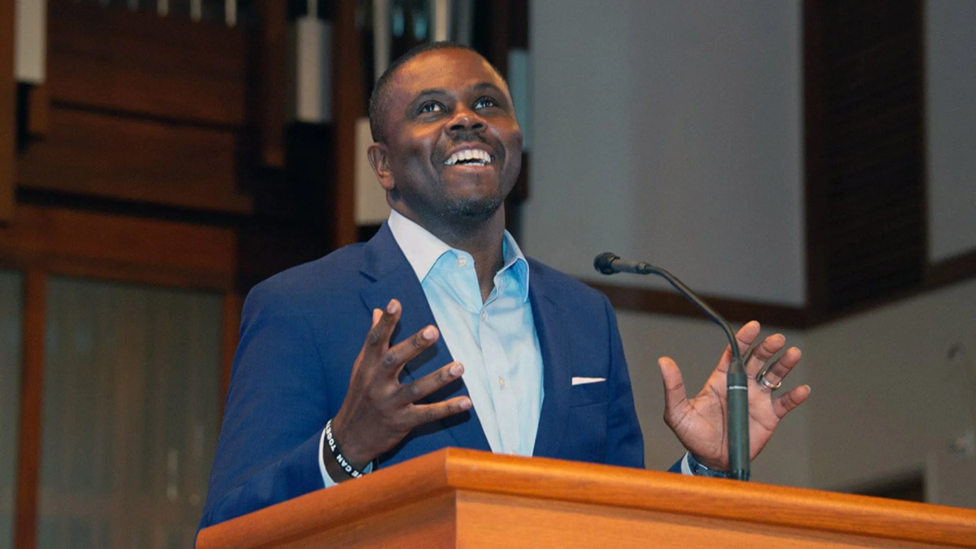 The Rev. Richie L. Butler, in a navy suit jacket and light blue shirt, smiles and looks upward with his hands raised as he speaks from a wooden pulpit with a microphone.