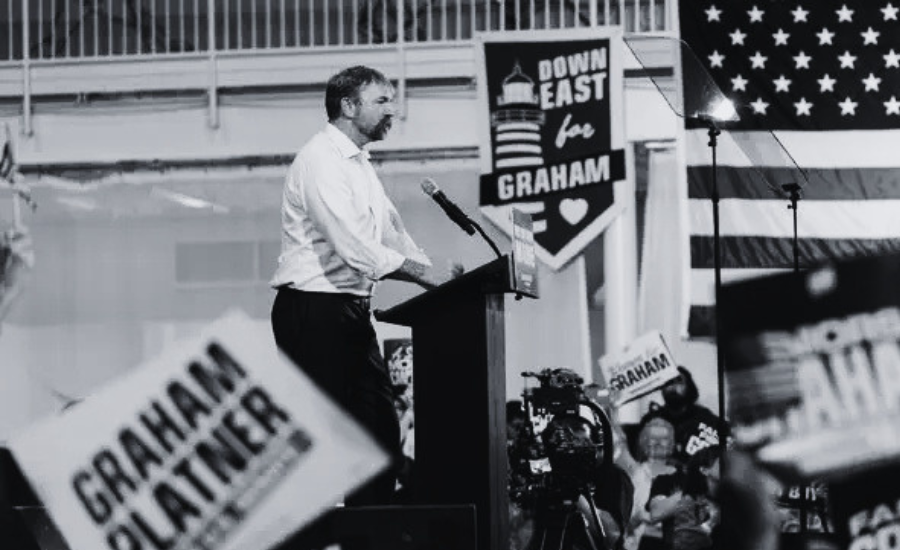 Graham Platner speaking at a podium at a Maine campaign rally, American flag and Graham Platner signs in background