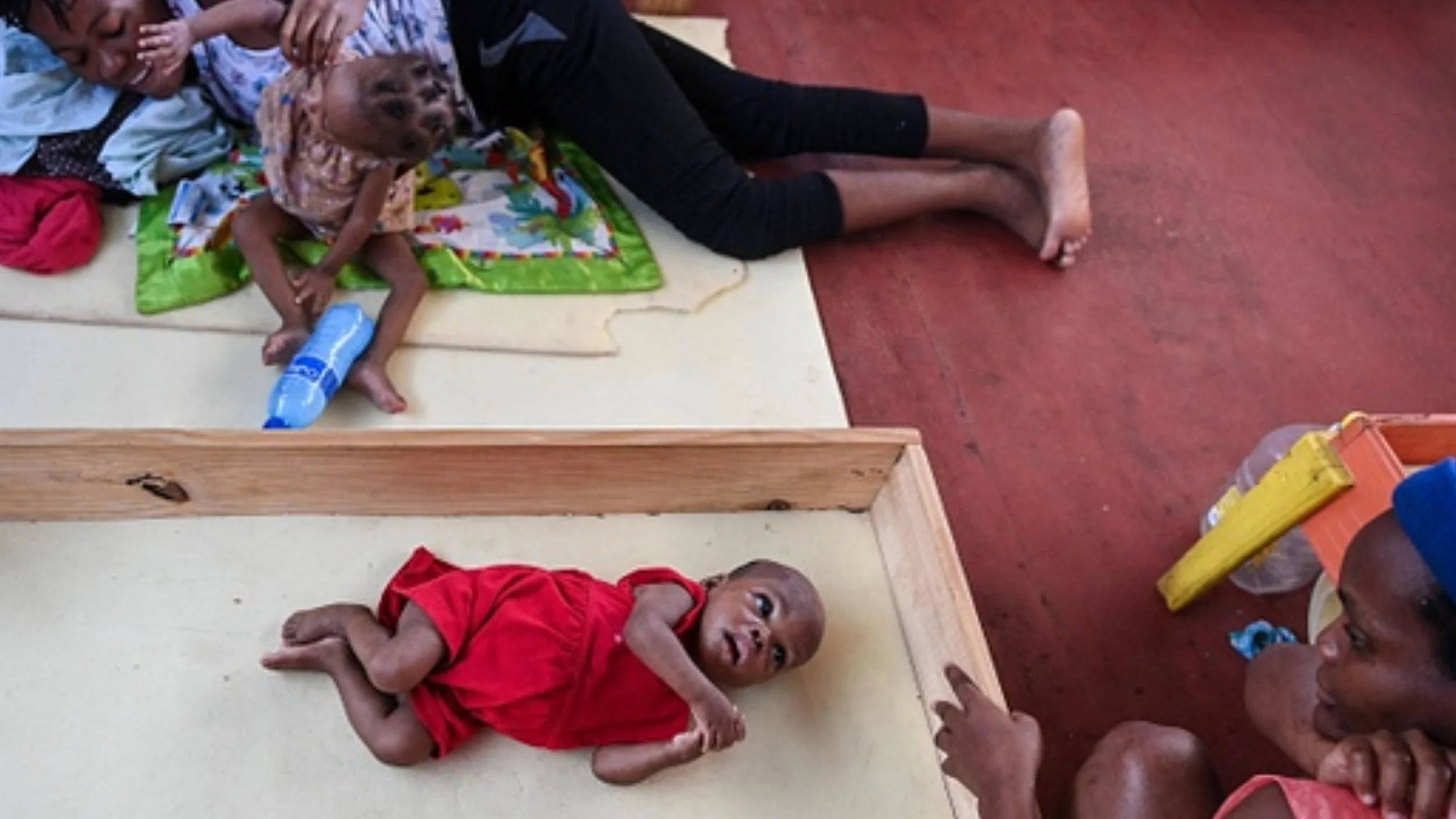 A thin baby in a red dress lies on a padded mat inside a wooden-framed bed as a young woman watches nearby, while a woman and a small child rest on a mat beside them.