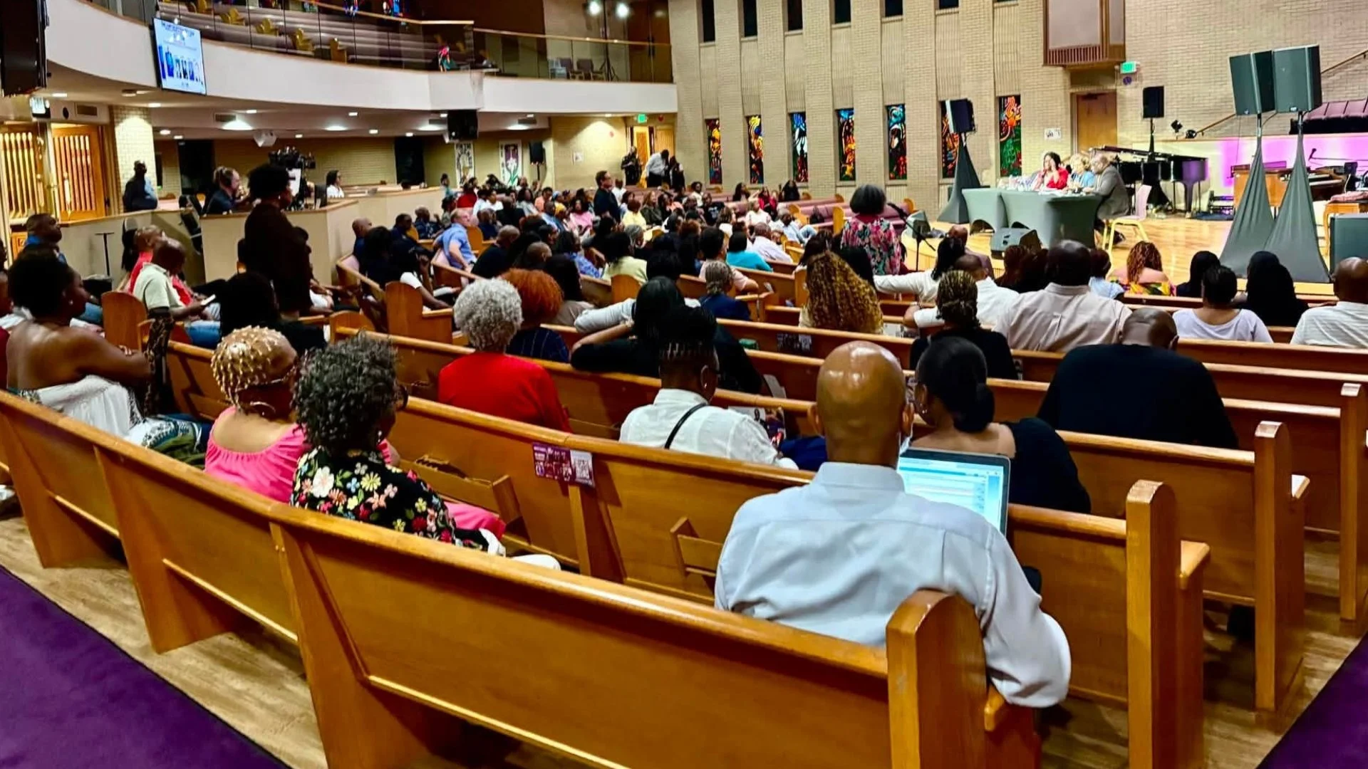 Rows of people seated in wooden pews inside a large church sanctuary with stained glass windows and purple carpet, facing a panel of speakers at a table at the front.