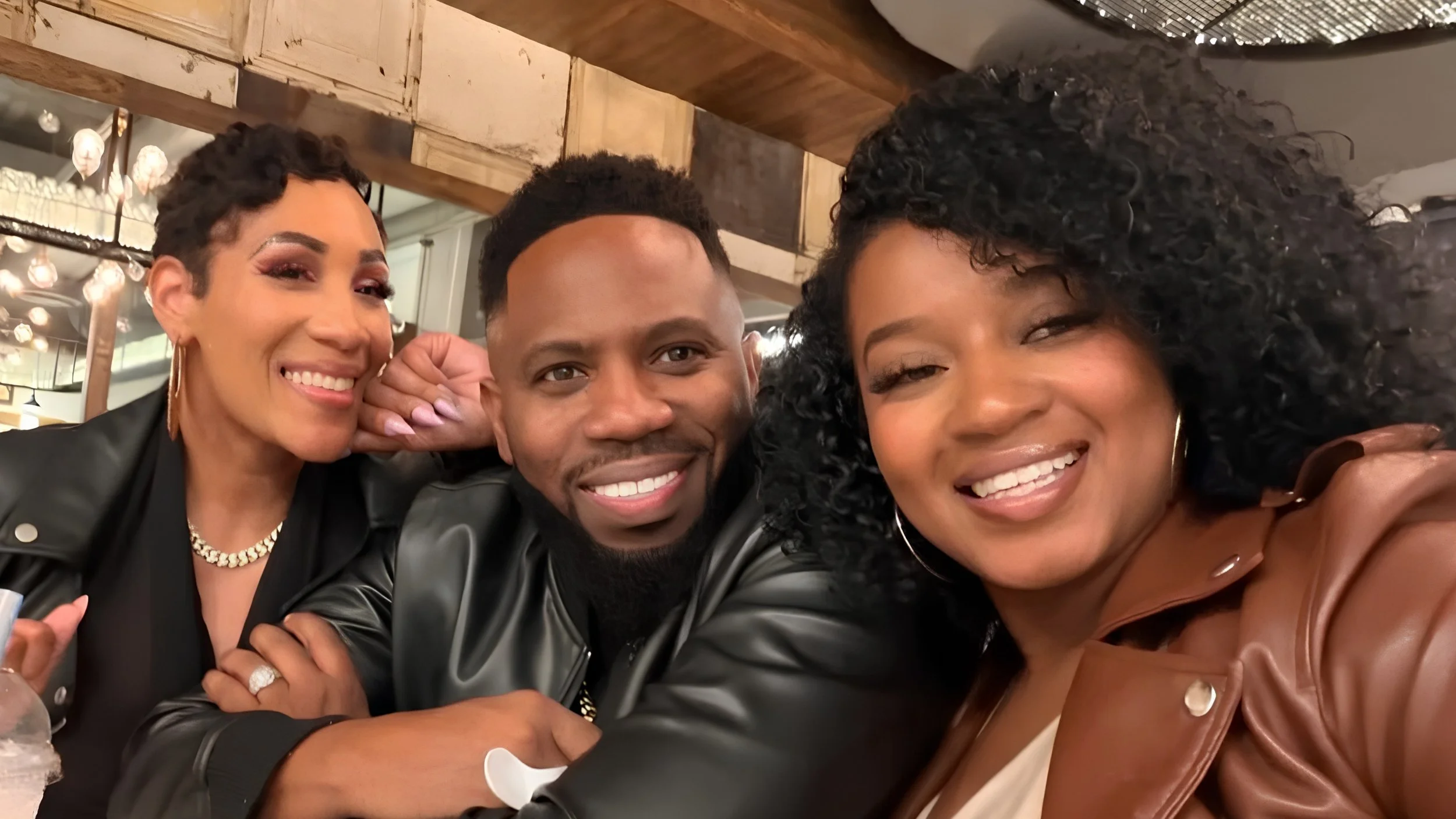 Israel Ford, in a black leather jacket, sits between his wives, Lydia Ford on the left and Shakayla Ford on the right, as the three smile for a selfie at a restaurant.