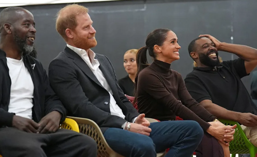 Harry and Meghan sit in a row of colorful plastic chairs between two smiling men, laughing as they watch a performance indoors.