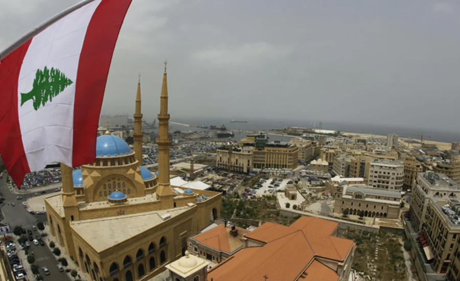 View of downtown Beirut, Lebanon, with a large Lebanese flag in the foreground and city buildings, including a mosque with blue domes and minarets, extending toward the Mediterranean coast in the distance.