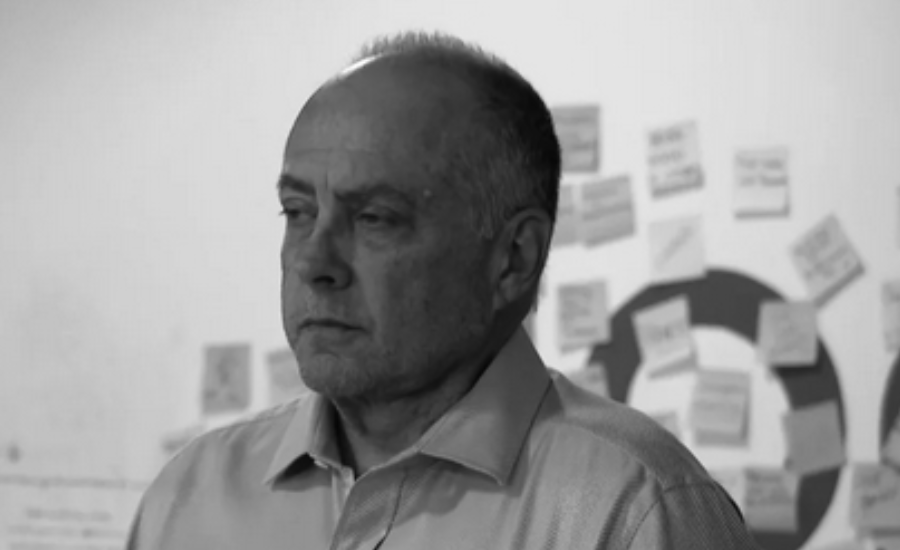 Black and white video screen grab of an older man with thinning gray hair and a serious expression, wearing a light collared shirt, looking off to the side. A wall covered with handwritten notes and papers is visible behind him.